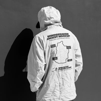 A person shown from behind wearing a white hooded jacket mockup with a large black printed graphic and text on the back, standing against a dark gray wall that casts a strong shadow.